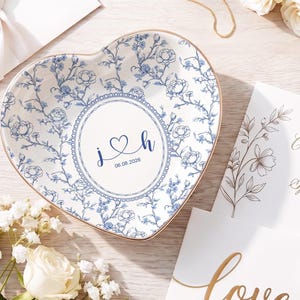 Puede incluir: Plato de cerámica en forma de corazón con diseño floral azul y borde dorado. El centro presenta las iniciales "J & H", un corazón y la fecha "06.08.2026". Tema de boda, con tarjetas y flores.