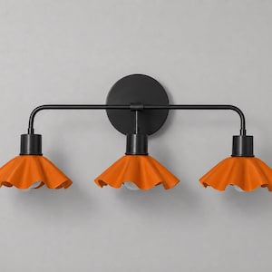 以下が含まれることがあります： マットブラックの金属製フレームと円形の壁掛け用ベースを備えた3灯式ウォールライト。鮮やかなオレンジ色のフリル付き円錐形シェードが3つ付いており、それぞれのシェードの下に電球ソケットが配置されています。