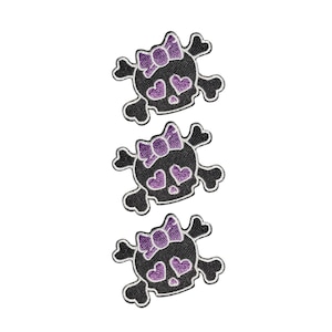 Puede incluir: Tres parches bordados idénticos con un diseño de calavera y huesos cruzados en negro. Cada calavera tiene un lazo morado en la parte superior y dos ojos en forma de corazón morados. El bordado está delineado con hilo blanco, creando contraste.