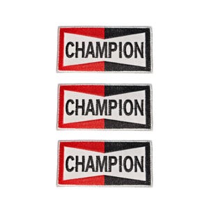 Op de afbeelding: Drie rechthoekige geborduurde patches met het woord CHAMPION in zwarte hoofdletters. Elke patch heeft een wit middengedeelte geflankeerd door een rode linkerkant en een zwarte rechterkant, allemaal omsloten door een dunne witte rand.