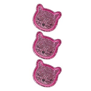 Puede incluir: Tres parches idénticos con forma de gato y acabado de purpurina rosa vibrante. Cada parche presenta un borde cosido, una pequeña nariz bordada en negro y bigotes negros bordados con los ojos cerrados en un diseño sencillo.