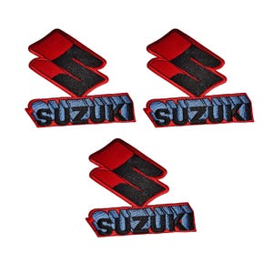 May include: Three embroidered patches featuring the Suzuki logo. Each patch has a red and black emblem above the word "SUZUKI" in blue and white lettering. The patches are on a white background.
