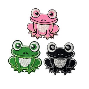 May include: Three embroidered frog patches in pink, green, and black. Each frog has large, round eyes and is smiling. The pink frog has a white belly, the green frog has a white belly, and the black frog has a white belly.