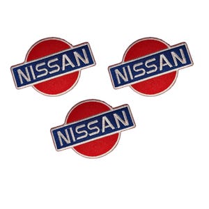 May include: Three embroidered Nissan logo patches. Each patch has a red circular background with a blue rectangular banner across it, displaying the white word "NISSAN". The patches are on a white background.