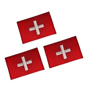 Puede incluir: Tres parches rectangulares rojos con una cruz blanca en el centro, que se asemejan a la bandera suiza. Los parches están dispuestos en diagonal sobre un fondo blanco. Los parches son probablemente bordados.