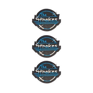Op de afbeelding: Drie identieke ronde geborduurde patches met een zwarte achtergrond en de tekst The Strokes in witte letters. Het ontwerp bevat blauwe cirkelvormige lijnen, witte accenten en een gestileerd kruisgrafiek, afgewerkt met een zwarte gestikte rand.