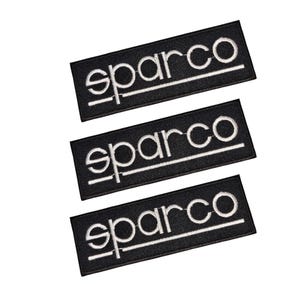 May include: Three black rectangular patches with the word "sparco" embroidered in white. The patches have a white horizontal line beneath the word. The patches are arranged diagonally against a white background.