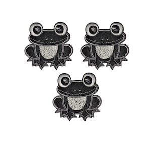 May include: Three embroidered frog patches. Each patch features a black frog with large, round eyes and a light grey belly. The patches are arranged in a triangular pattern against a white background. The frog's legs are a dark grey colour.
