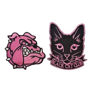 Peut inclure: Deux écussons brodés : un bouledogue rose avec des pointes blanches et une tête de chat noire avec le texte "BLACK CAT CLUB" en rose. Les écussons sont pour vêtements ou accessoires.