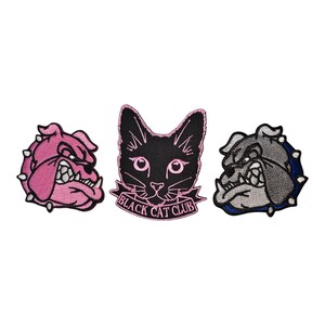 Può includere: Tre toppe ricamate: un bulldog rosa, un gatto nero con la scritta "BLACK CAT CLUB" e un bulldog grigio. Le toppe del bulldog hanno un'espressione feroce. La toppa del gatto ha accenti rosa.