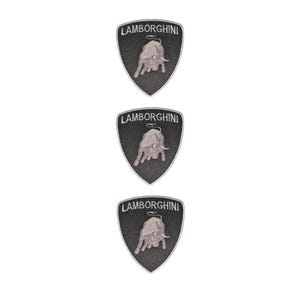 Puede incluir: Tres parches bordados idénticos en forma de escudo con el logotipo de Lamborghini. Cada parche tiene un fondo negro con un emblema de toro de color plateado en el centro y la palabra LAMBORGHINI bordada en letras blancas en la parte superior.