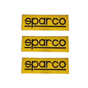 Puede incluir: Tres parches rectangulares amarillos con la palabra "sparco" en negro, centrada. El texto está encima de una línea negra horizontal. Los parches son para ropa de automoción o de carreras.