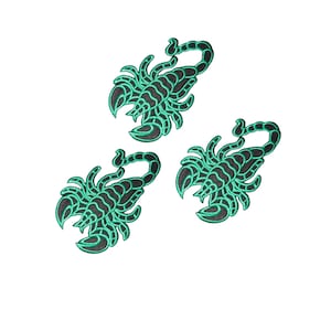 Könnte beinhalten: Drei gestickte Skorpion-Aufnäher mit schwarzer Basis und leuchtend türkisfarbenen Nähten, die den segmentierten Körper, die Scheren und den gebogenen Schwanz hervorheben. Jeder Aufnäher zeigt ein detailliertes Insektendesign mit erhabener Struktur.
