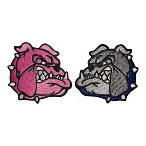 May include: Two embroidered patches featuring bulldog faces. One is pink with white accents, the other is gray with blue accents. Both have a fierce expression with exposed teeth and white spikes around the edges.