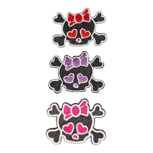 Puede incluir: Tres parches bordados con calaveras negras y huesos cruzados. Cada calavera tiene ojos en forma de corazón y una pequeña nariz. El parche superior tiene detalles rojos, el central morados y el inferior rosas, todos con lazos decorativos.