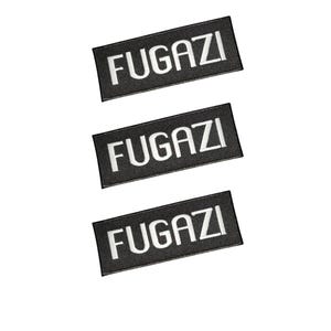 Puede incluir: Tres parches rectangulares negros dispuestos verticalmente, cada uno con la palabra FUGAZI bordada en letras mayúsculas blancas y negritas. Los parches tienen un borde cosido limpio y un diseño plano uniforme para aplicar sobre diversas telas.