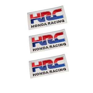 Könnte beinhalten: Drei rechteckige gestickte Aufnäher mit dem HRC-Logo in rotem und blauem Garn auf weißem Hintergrund. Unter dem HRC-Schriftzug ist auf jedem der drei identischen Aufnäher der Text HONDA RACING in schwarzem Garn eingestickt.