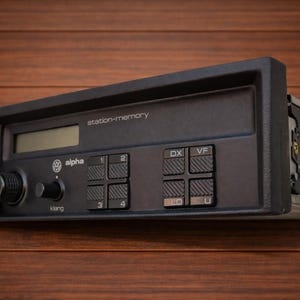 Può includere: Un'autoradio Volkswagen Alpha nera dotata di display digitale, manopola del volume, manopola di controllo del tono etichettata Klang e otto pulsanti testurizzati. Il pannello frontale include il testo station-memory e il logo Volkswagen.