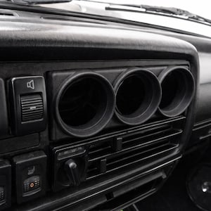 May include: A black automotive dashboard interior featuring three circular gauge pods positioned above horizontal air conditioning vents. The console includes various rectangular push buttons with white icons, a volume slider, and seat heater controls.