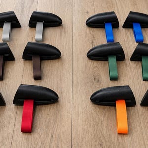 Könnte beinhalten: Zwölf schwarze Knebelverschlüsse, paarweise angeordnet, jeweils mit einer farbigen Schlaufe. Die Schlaufen sind in Silber, Braun, Rot, Blau, Grün und Orange gehalten. Die Verschlüsse bestehen aus einem gebogenen schwarzen Knebel und einer Stoffschlaufe.