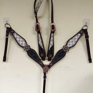 May include: A dark brown leather horse breast collar featuring a checkered pattern in light gray and white, accented with black leather sections, copper-toned metal buckles, floral-engraved hardware, and circular silver-toned stud embellishments.