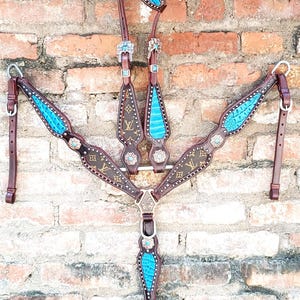 May include: A brown leather horse breast collar and headstall set featuring dark brown panels with a repeating gold monogram pattern, accented by bright turquoise textured inlays, silver-toned metal hardware, and decorative floral conchos.