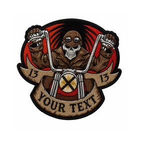 May include: A circular embroidered patch featuring a skull wearing goggles and a brown jacket, riding a motorcycle with handlebars visible. A tan banner across the bottom displays the text YOUR TEXT, with the number 13 on each side against a red background.