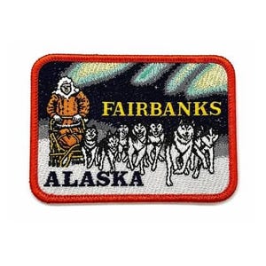 Pode incluir: Um emblema bordado retangular com borda vermelha que mostra uma equipe de trenó puxada por cães, guiada por um musher com parka. O design inclui o texto FAIRBANKS em amarelo e ALASKA em preto sobre um fundo escuro com auroras boreais verdes.