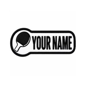 May include: A black and white patch featuring a ping pong paddle graphic next to the text "YOUR NAME". The patch has a black border and a white background. The ping pong paddle is black with a white handle.