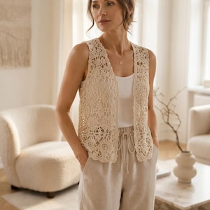 Può includere: Un gilet senza maniche all'uncinetto color crema caratterizzato da un intricato motivo floreale con bordi smerlati. Il gilet è indossato sopra una semplice canotta bianca a coste e abbinato a pantaloni di lino beige chiaro con coulisse.