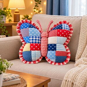 Può includere: Un cuscino a forma di farfalla caratterizzato da un design patchwork con quadrati di tessuto rossi, blu, bianchi e gialli. I motivi includono pois, quadretti vichy e colori a tinta unita. Il cuscino presenta un bordo arricciato a quadretti.
