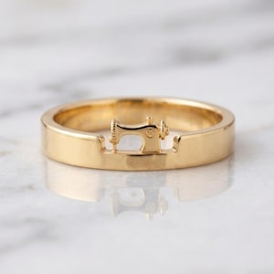 May include: A polished gold band ring featuring a small, integrated three-dimensional sewing machine motif centered on the top. The ring has a smooth, reflective surface and a uniform width, showcasing a detailed miniature sewing machine design.