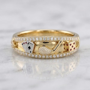 May include: A gold ring featuring two rows of small clear stones along the edges. The center band displays a miniature set of playing cards, a textured golf club, and a gold die with red dots. The design incorporates symbols of luck and leisure.