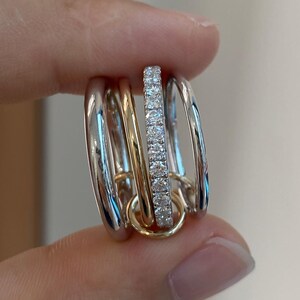 May include: A multi-band ring featuring a central band of small, sparkling stones, flanked by bands of polished gold and silver. The ring is held between fingers, showcasing its intricate design and mixed metal composition.