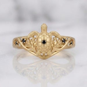 Op de afbeelding: Een gouden ring met een centraal zeeschildpadmotief met een kleine zwarte edelsteen in het schild. De band is ontworpen met ingewikkeld, takachtig metaalwerk dat zich uitstrekt vanaf de schildpad, geaccentueerd door kleine zwarte en heldere edelstenen.
