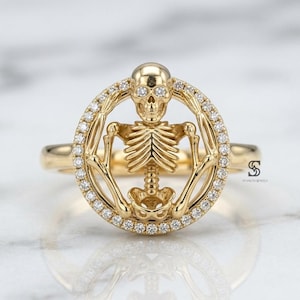 Könnte beinhalten: Ein goldener Ring mit einem kreisförmigen Rahmen, der mit kleinen weißen Steinen besetzt ist. Im Inneren des Kreises befindet sich eine detaillierte goldene Skelettfigur. Die Ringschiene ist glatt und poliert. Ein kleines Logo mit SD ist sichtbar.
