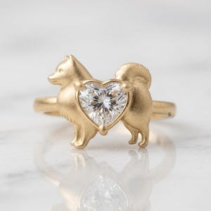 May include: A gold ring featuring a matte finish dog silhouette with a clear, faceted heart-shaped gemstone set in the center. The dog figure is depicted in profile, and the band is a simple, smooth gold hoop that encircles the finger.