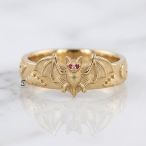 May include: A gold ring featuring a central bat motif with two small, vibrant pink gemstone eyes. The band is detailed with embossed cloud and crescent moon shapes on either side of the bat, creating a continuous, textured design around the ring.