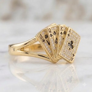 May include: A gold ring featuring a fan of five playing cards as the centerpiece. Each card is encrusted with small clear stones and displays the symbols for a royal flush in clubs, including the Jack, Queen, King, and Ace of clubs.