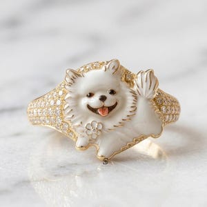 May include: A gold ring featuring a white enamel Pomeranian dog design with a small flower accent. The band and the dog's body are encrusted with numerous small, clear, sparkling stones. The dog has a black nose, black eyes, and a pink tongue.