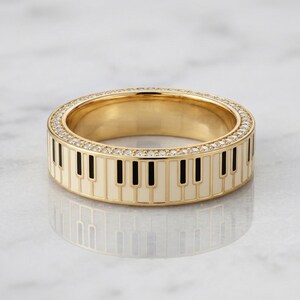 May include: A gold band ring featuring a repeating piano key pattern in black and cream enamel around the exterior. The top and bottom edges of the ring are lined with a continuous row of small, clear, sparkling stones set in gold metal.