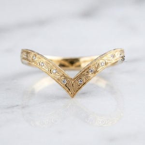 Puede incluir: Un anillo de oro en forma de V con intrincados patrones de volutas grabados a lo largo de la banda. El anillo está acentuado con pequeñas piedras redondas y transparentes engastadas a intervalos en el metal detallado.