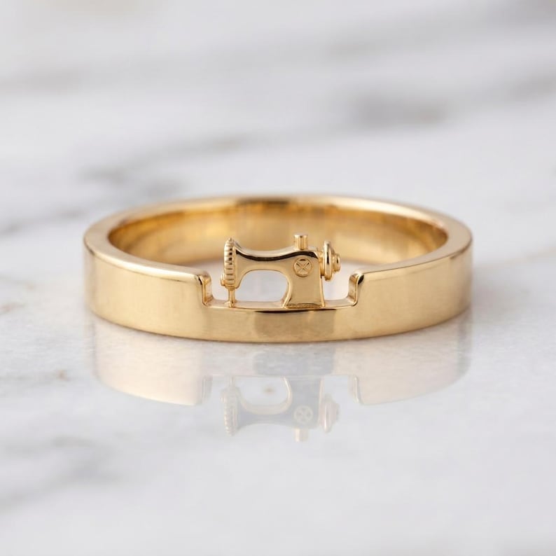 May include: A polished gold band ring featuring a small, integrated three-dimensional sewing machine motif centered on the top. The ring has a smooth, reflective surface and a uniform width, showcasing a detailed miniature sewing machine design.