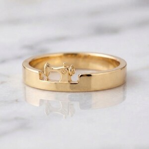 May include: A polished gold ring featuring a small, integrated three-dimensional sewing machine motif centered within a recessed section of the band. The metal surface is smooth and reflective, showcasing a continuous circular design.
