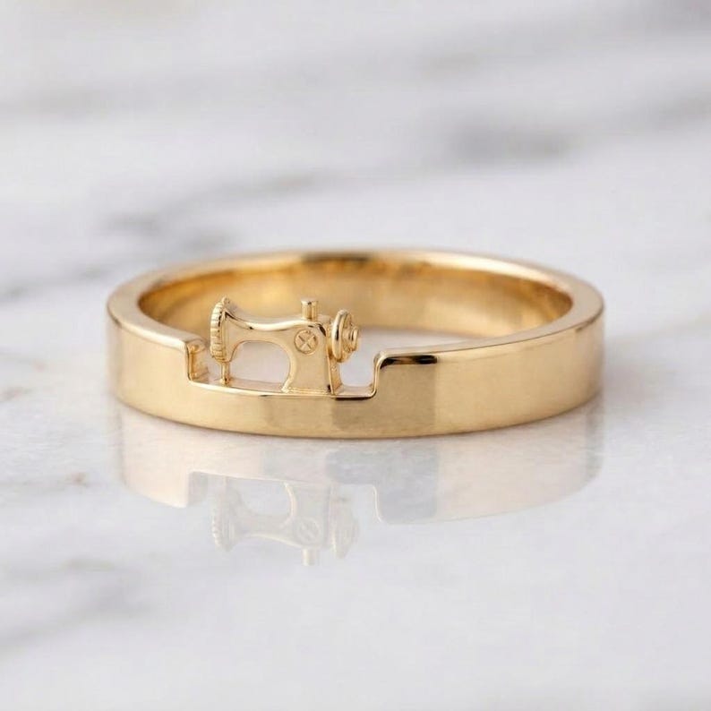May include: A polished gold ring featuring a small, integrated three-dimensional sewing machine motif centered within a recessed section of the band. The metal surface is smooth and reflective, showcasing a continuous circular design.