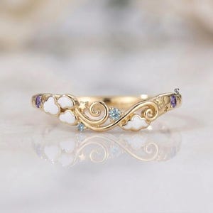 May include: A gold band ring featuring white enamel cloud motifs, small engraved stars, and inlaid gemstones including light blue and purple stones. The design incorporates swirling metal patterns and a curved shape that follows a celestial theme.