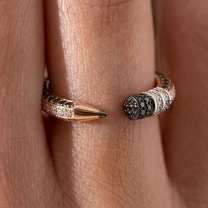 May include: An open-style rose gold ring shaped like a pencil, featuring a pointed tip with black and white pavé stones on one end and a flat eraser-like end with black and white pavé stones on the other, creating a continuous wrap-around design.