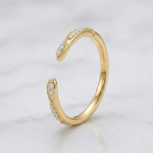 May include: A gold-colored open ring with a curved design. The ring is adorned with small, sparkling diamonds along the curved edges. The ring is set against a white background.