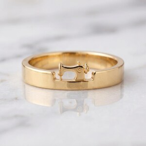 May include: A polished gold ring featuring a small, detailed three-dimensional sewing machine motif integrated into the center of the band. The metal has a smooth, reflective finish, and the design showcases the distinct shape of a vintage sewing machine.
