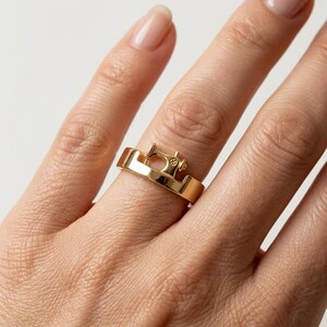May include: A polished gold-tone ring featuring a small, detailed three-dimensional sewing machine motif integrated into the band. The metal surface is smooth and reflective, showcasing the distinct shape of the sewing machine mechanism.